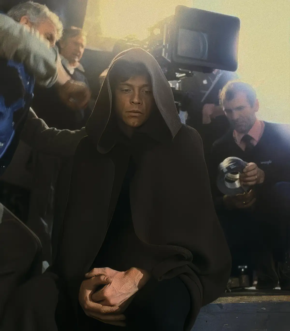 A man in a dark hooded robe sits dejectedly in what appears to be a film set, surrounded by crew members and equipment. The…