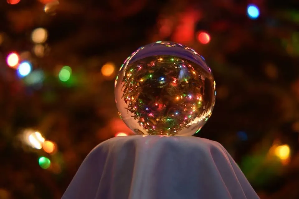 A clear glass crystal ball sits atop a white cloth-covered stand, reflecting a blurred background of colorful Christmas tree…