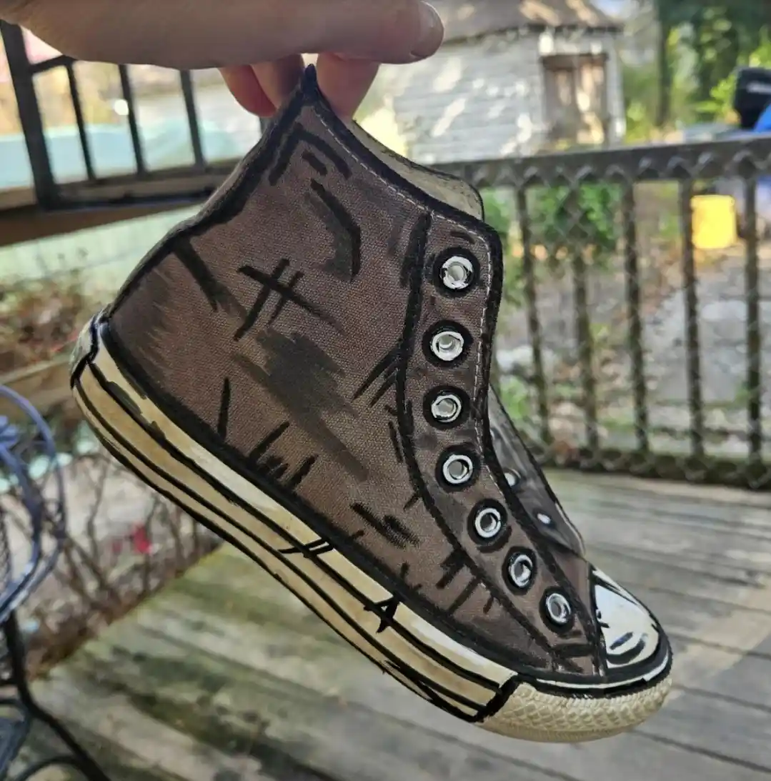 A hand holds up a high-top sneaker that has been painted to look like a drawing of a shoe, which itself is painted to look…