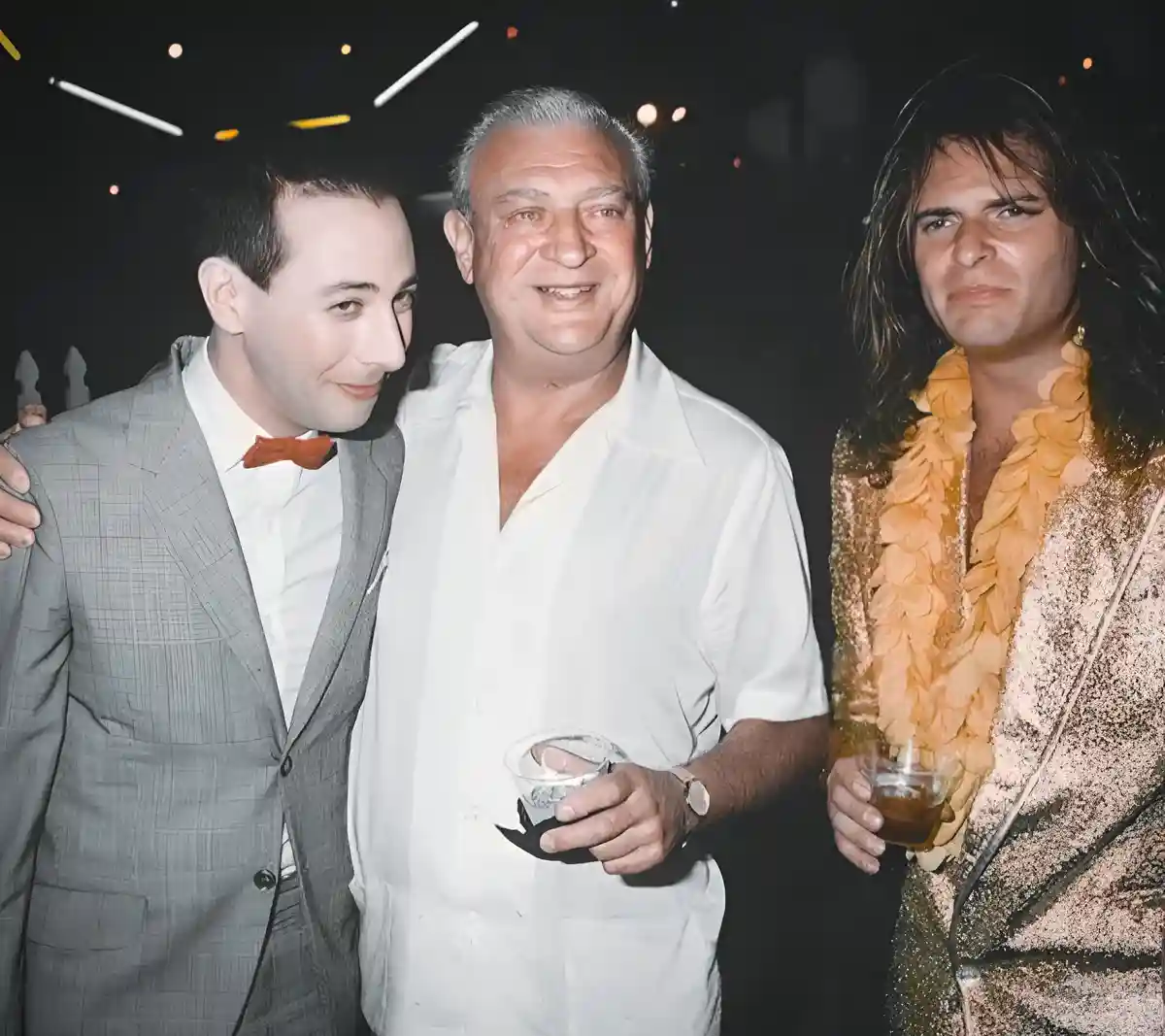 Three men pose together at a party; the man on the left in a suit and bowtie smiles while looking at the center man, who is…