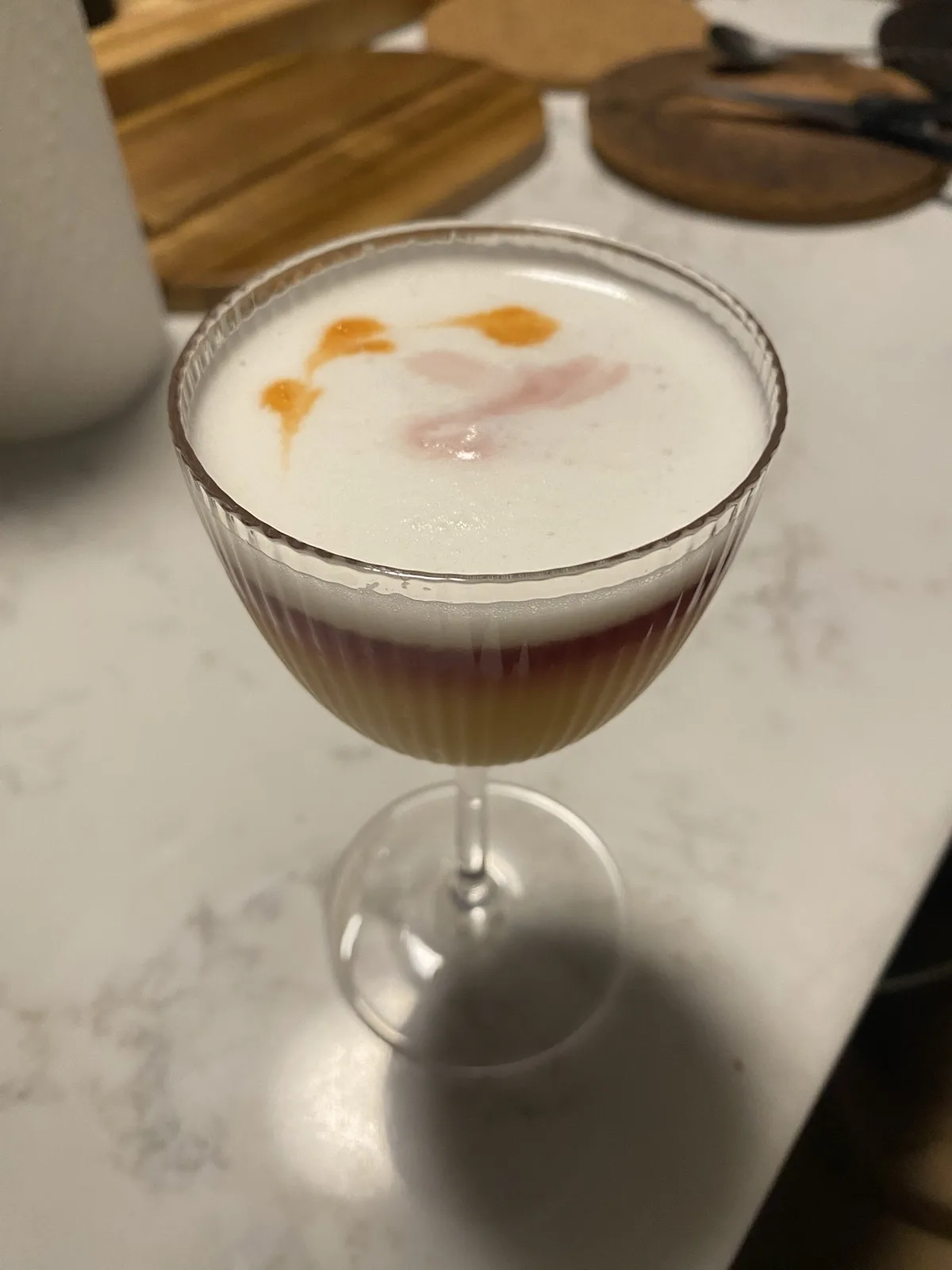 A close-up photo of a cocktail in a ribbed coupe glass, featuring a white frothy top with orange and pink swirls that…