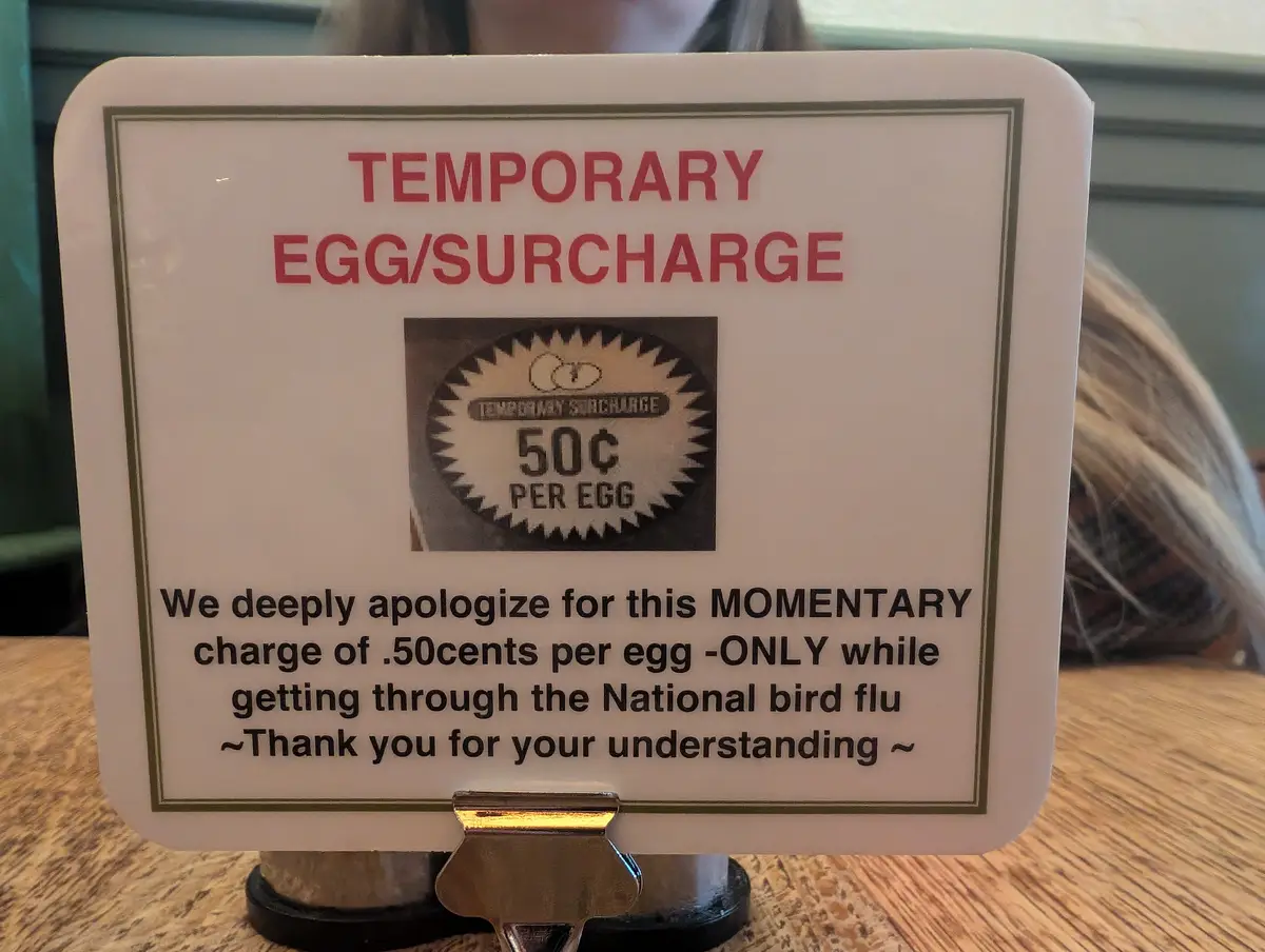 A white sign on a table announces a 'TEMPORARY EGG/SURCHARGE' of 50 cents per egg, with a small graphic of the charge. The…