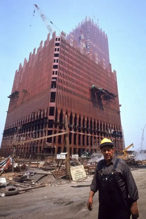 A construction worker in overalls and a yellow hard hat stands in front of a partially built skyscraper with a red steel…