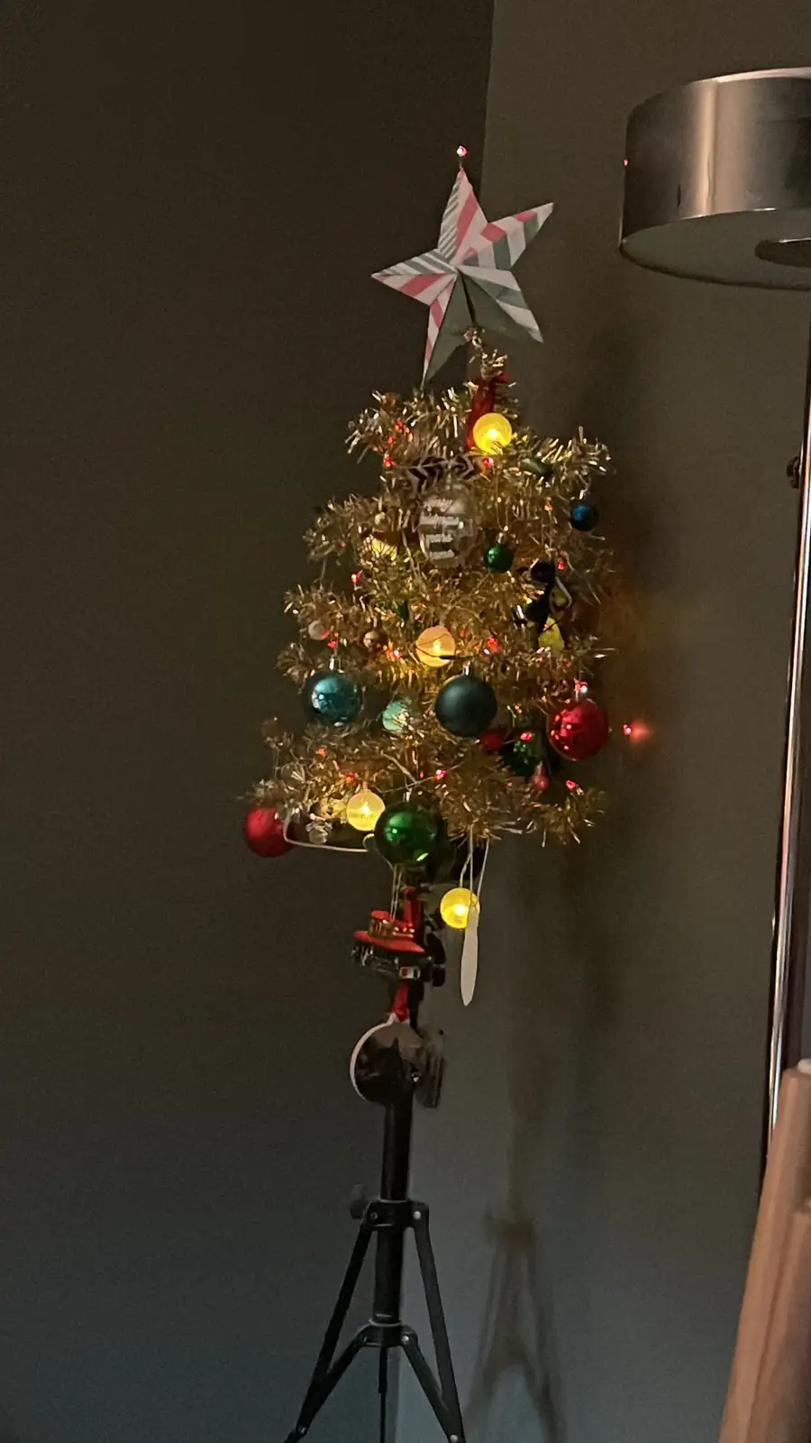 A small, gold tinsel Christmas tree decorated with colorful ornaments, lights, and a striped star topper is mounted on a…