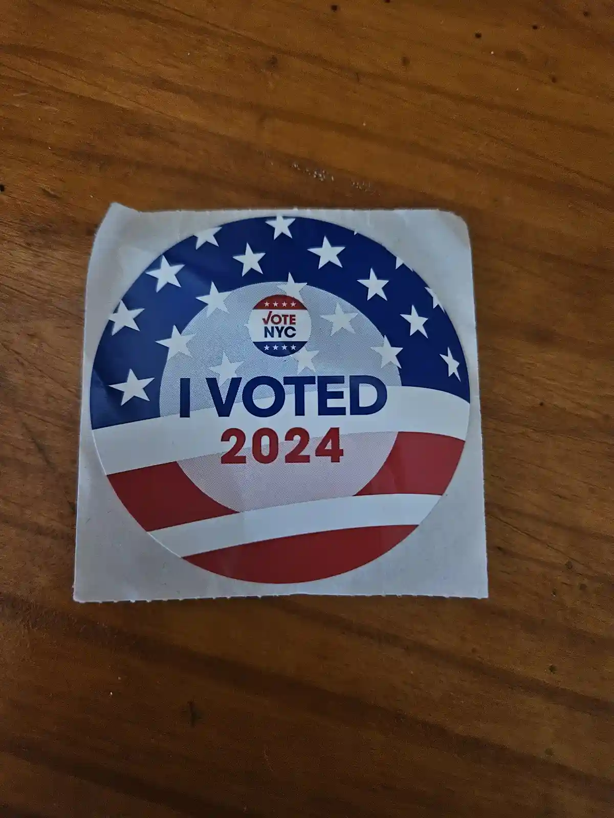 A circular 'I VOTED 2024' sticker with American flag design (stars and stripes) and 'VOTE NYC' logo, placed on a wooden…