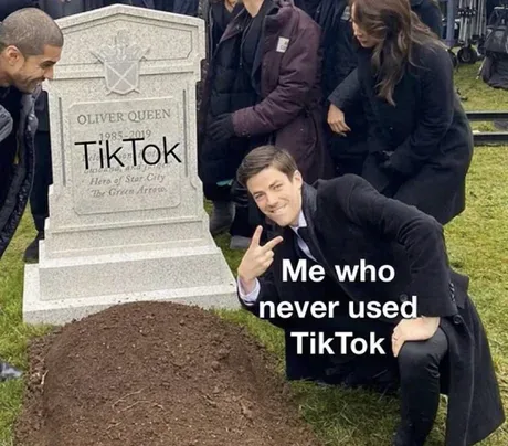 A man in a black suit poses cheerfully in front of a tombstone labeled 'Oliver Queen 1985-2019 Hero of Star City The Green…