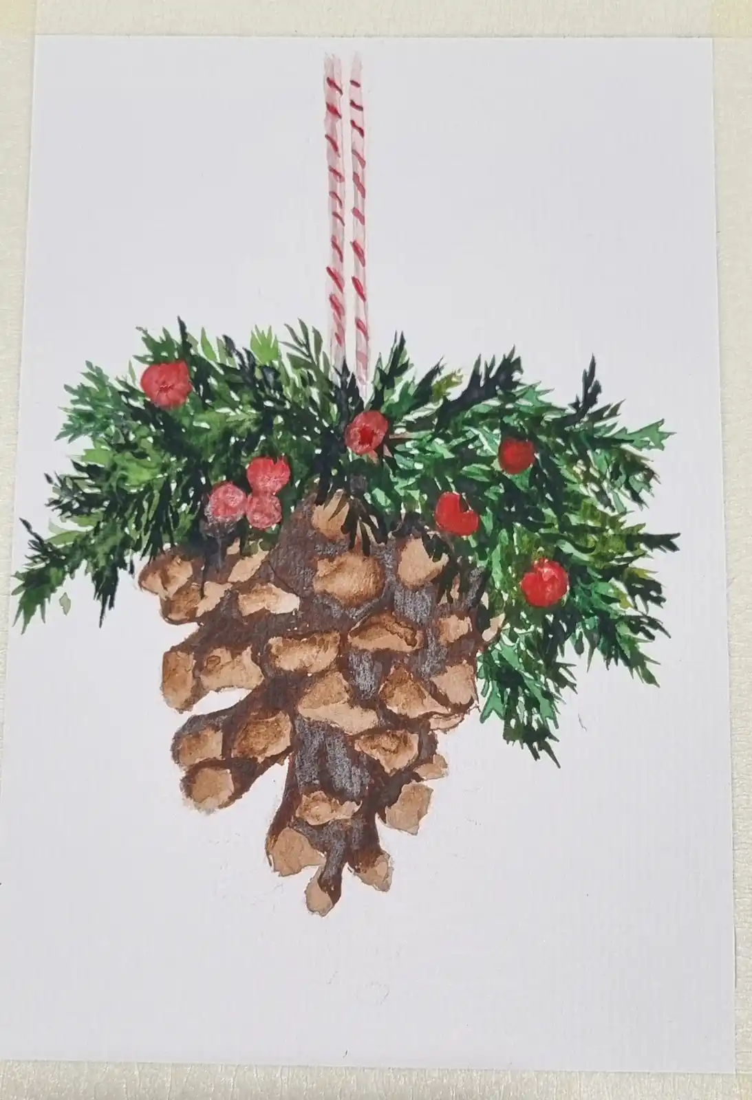 A watercolor painting of a pinecone decorated with green pine needles and red berries, hanging from two red-and-white…