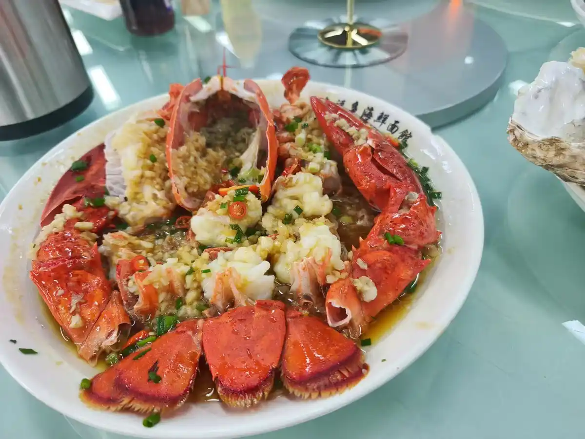 a close-up photo shows a large white plate filled with steamed lobster claws and tail sections, generously topped with…