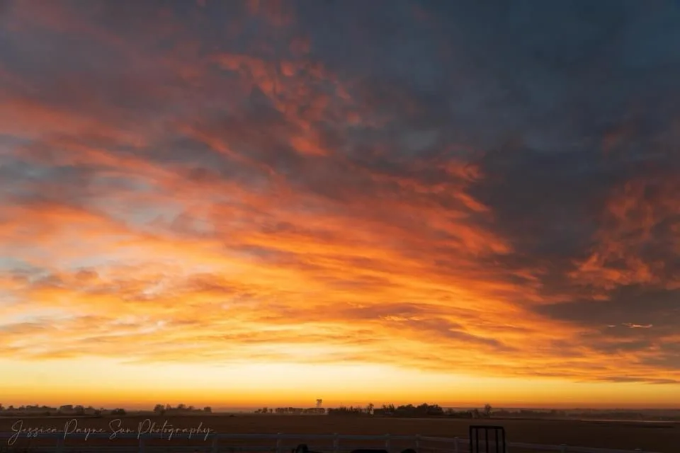 A dramatic sunrise or sunset photo showing fiery orange and red clouds over a flat horizon with a white fence in the…