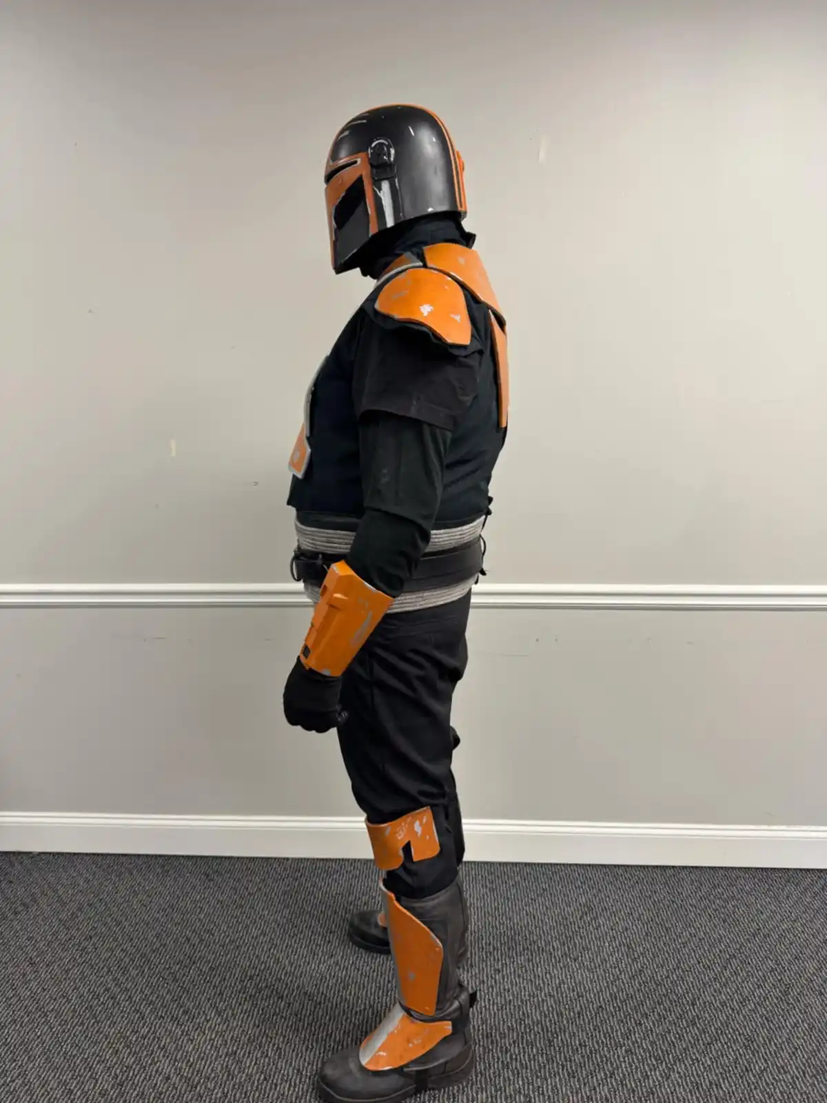 A person in a full-body Mandalorian armor costume, featuring black and orange plating, stands in profile against a plain…