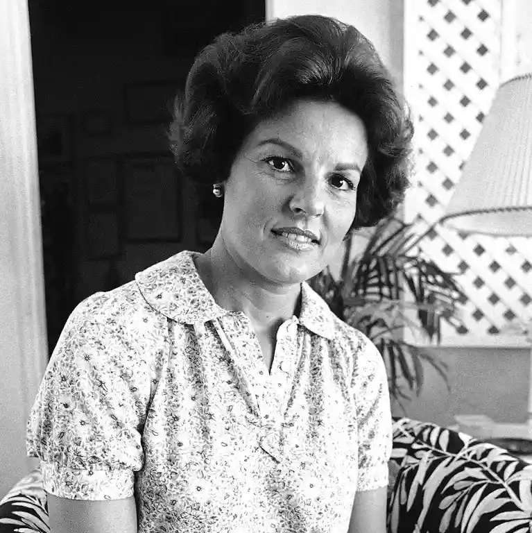 A black-and-white photo of a woman with styled hair, wearing a floral-patterned blouse, seated indoors with a lamp and…