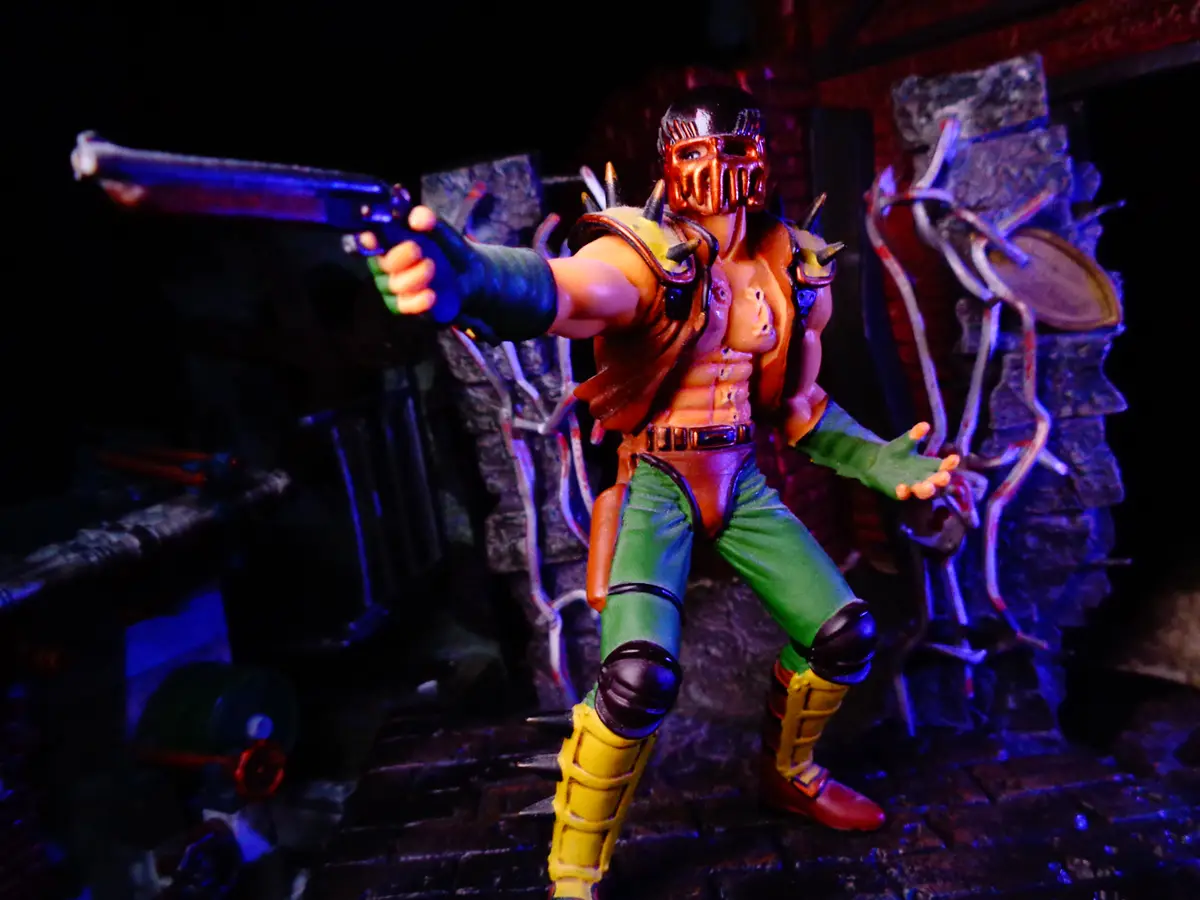 A toy figure of the video game character Reptile from Mortal Kombat, posed with a pistol aimed forward, stands in a dark…