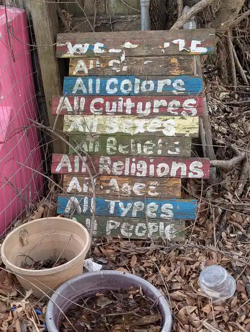 A weathered, multi-colored wooden sign made of stacked planks stands in a leafy, overgrown yard next to a pink fence and…
