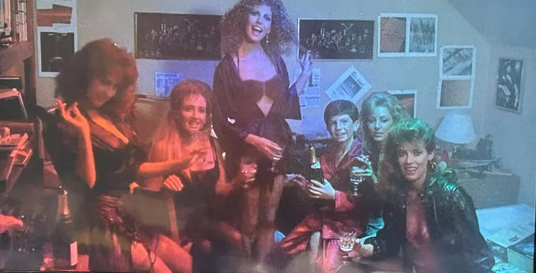 A group of six people, mostly women, are gathered in a dimly lit room with posters on the wall, holding drinks and posing…