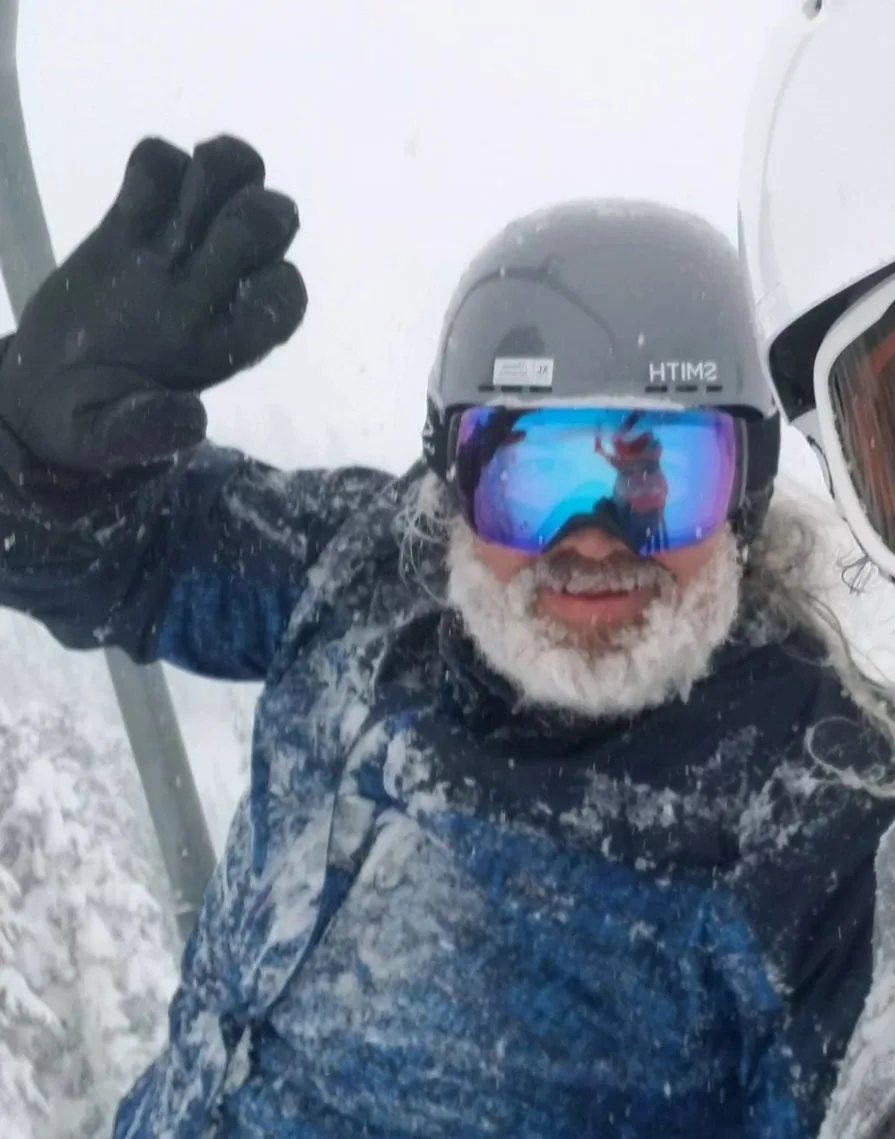 A person in full winter gear, including a helmet and goggles, sits on a ski lift covered in snow. Their beard is frosted…
