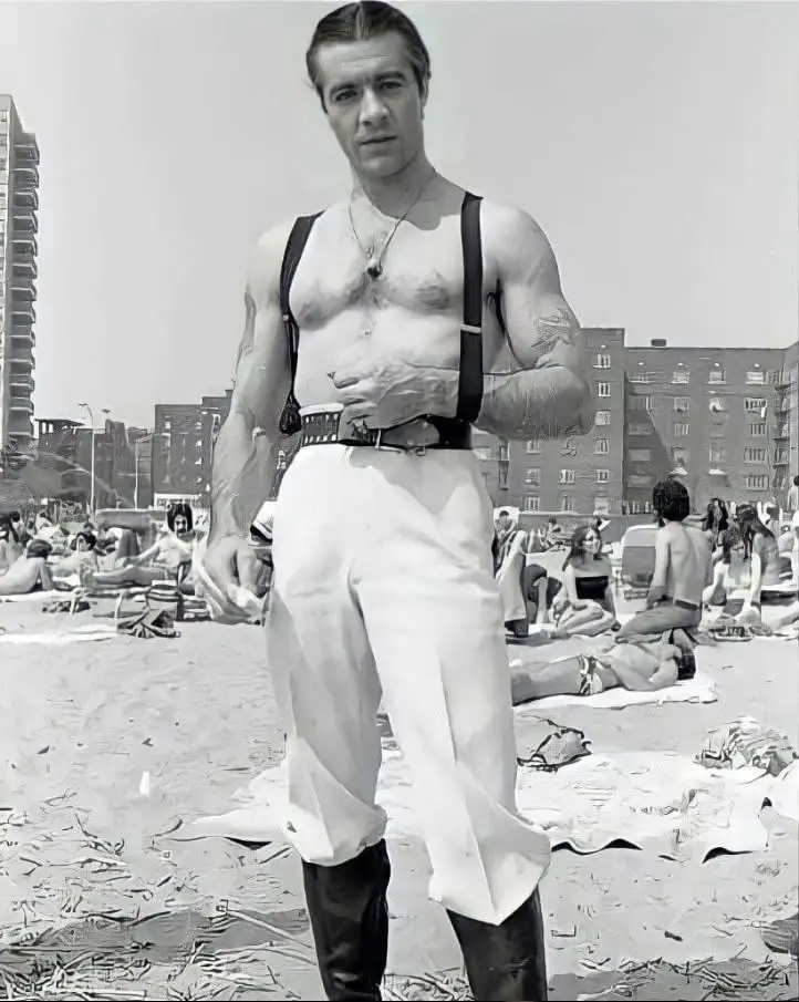 A black-and-white photo shows a muscular man standing on a crowded beach, wearing white pants held up by suspenders, black…