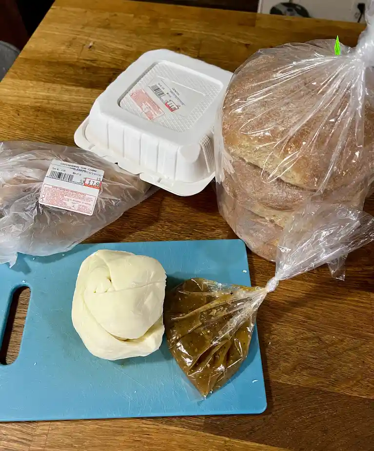 A wooden table holds groceries: a white takeout container, a large plastic bag of bread rolls, a plastic-wrapped package of…