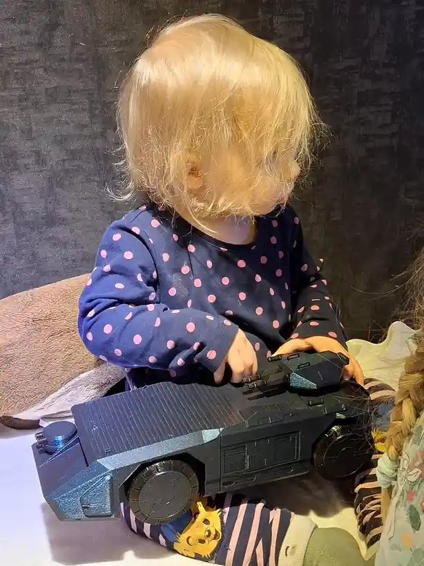 A toddler with blonde hair wearing a blue shirt with pink polka dots is sitting on the floor and holding a large black toy…