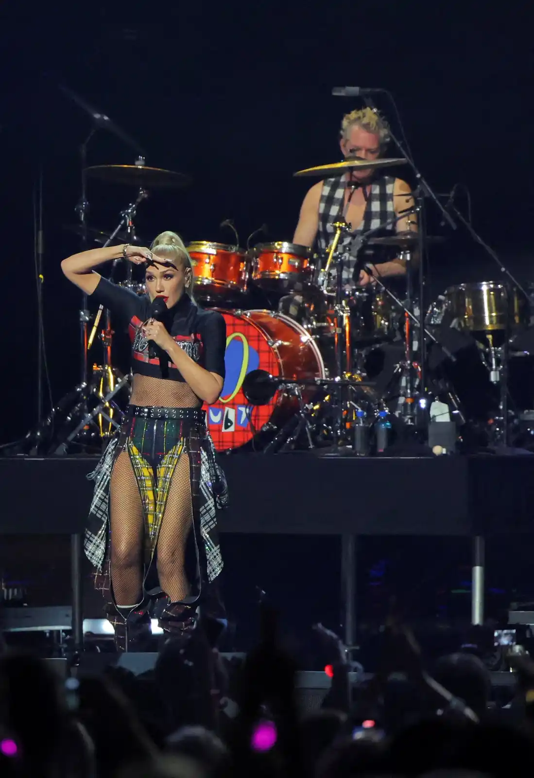 A woman with blonde hair in a high ponytail, wearing a black crop top, fishnet shorts with a plaid skirt overlay, and high…