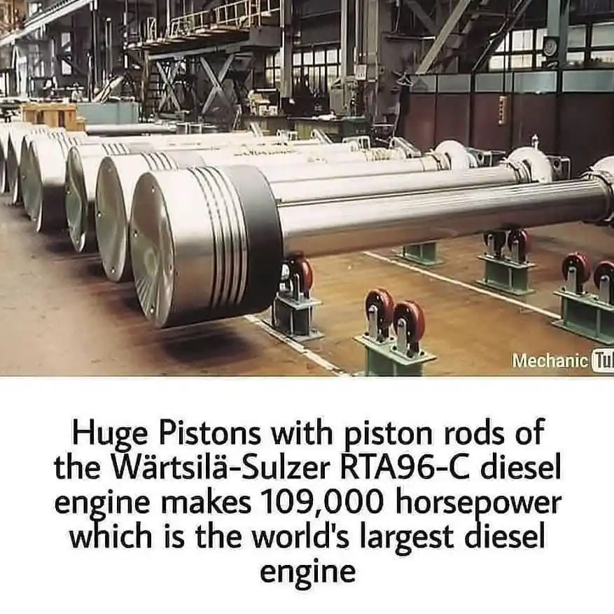The image shows a row of massive, cylindrical metal pistons with attached piston rods, resting on wheeled stands inside a…