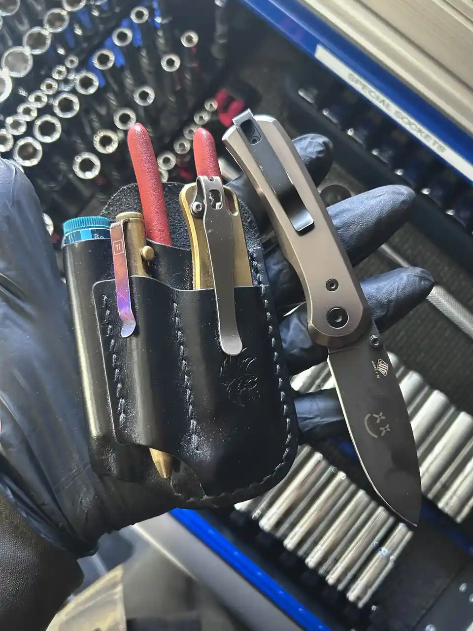 A hand in a black glove holds a black leather tool pouch containing various small tools like a lighter, tweezers, and a…