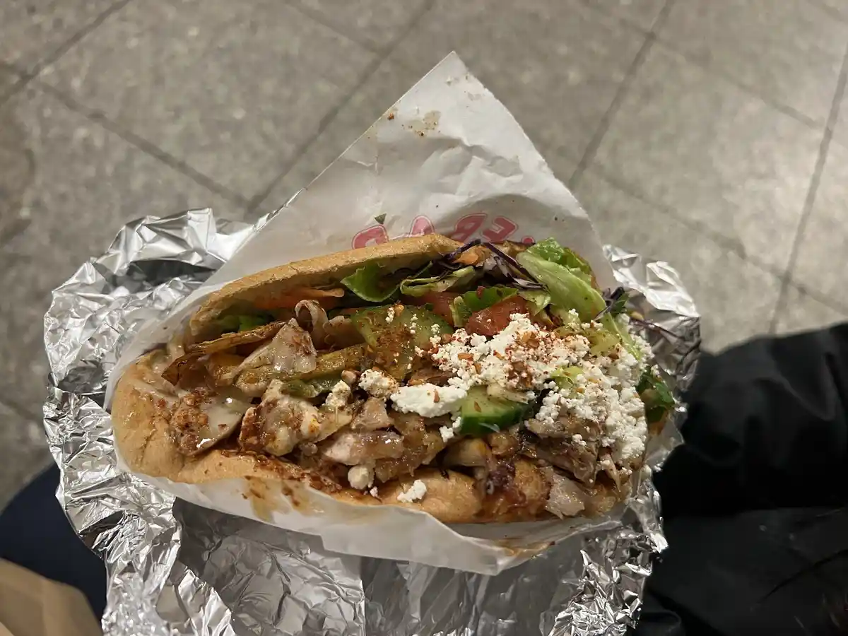 A close-up photo shows a generously filled chicken gyro sandwich held in foil, topped with lettuce, tomato, cucumber…