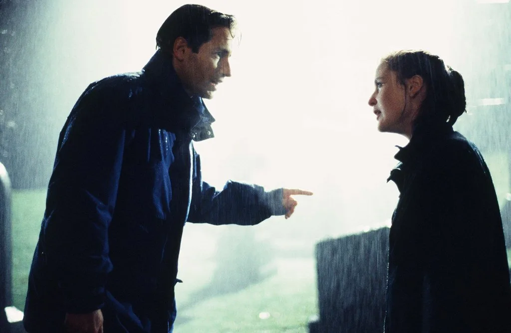 a man in a dark jacket points accusingly at a woman in a dark jacket standing in heavy rain, both looking intensely at each…