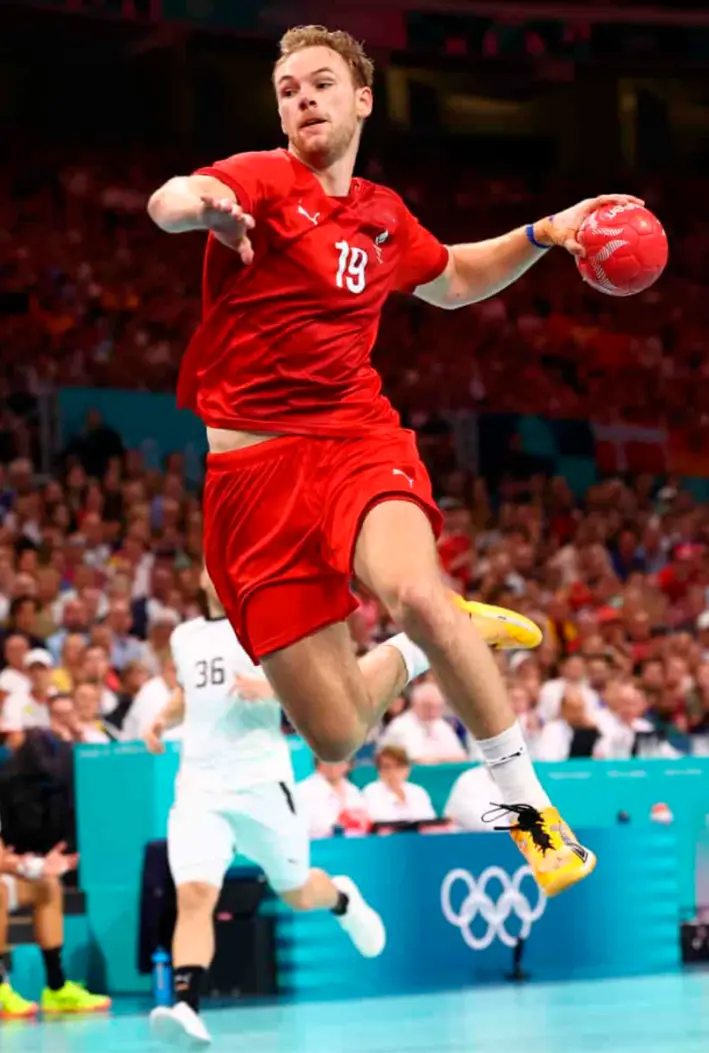 A male handball player in a red jersey with the number 19 is mid-air, holding a red handball, wearing bright yellow shoes…
