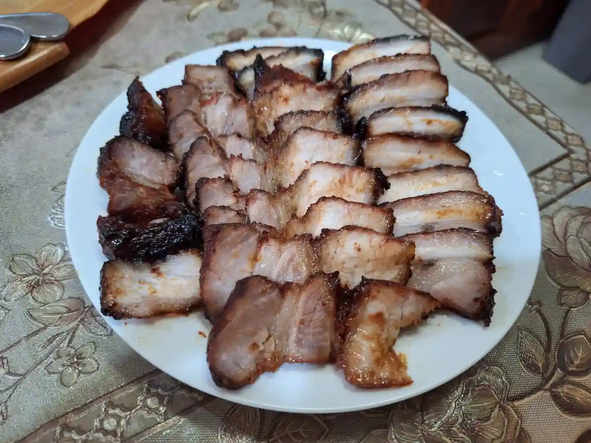 A white plate holds neatly arranged slices of roasted pork with a caramelized, slightly charred exterior, resting on a…