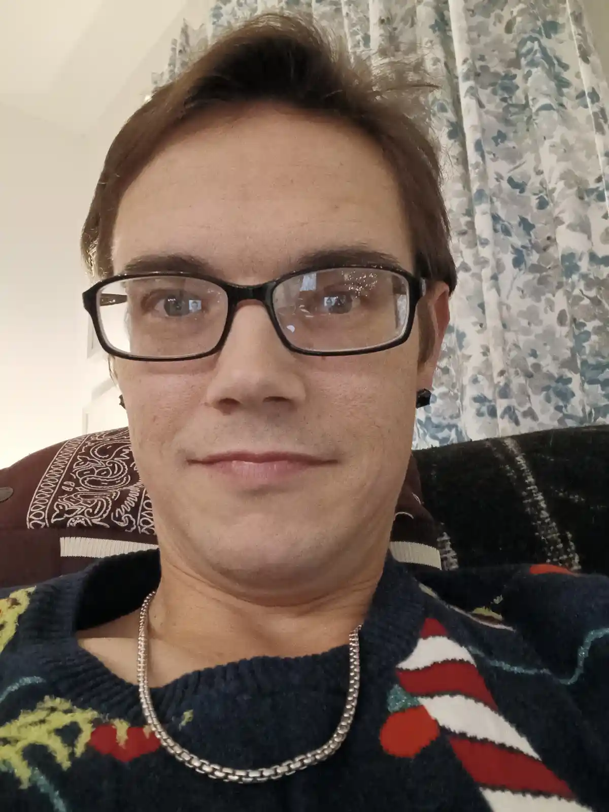 A close-up selfie of a man with brown hair and black-rimmed glasses, wearing a colorful patterned sweater and a silver chain…