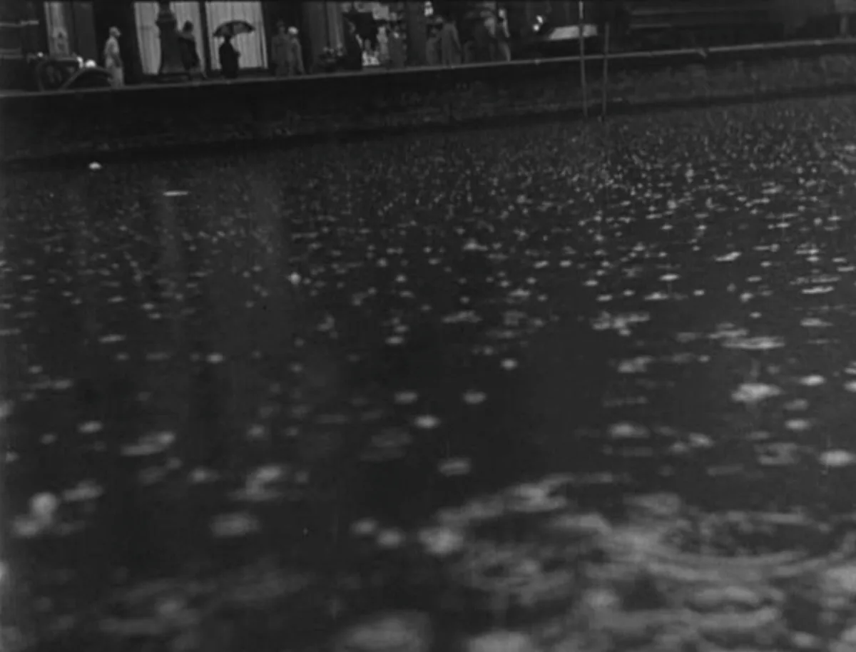 A black-and-white historical photograph shows raindrops hitting a wet street, creating ripples and reflections. In the…