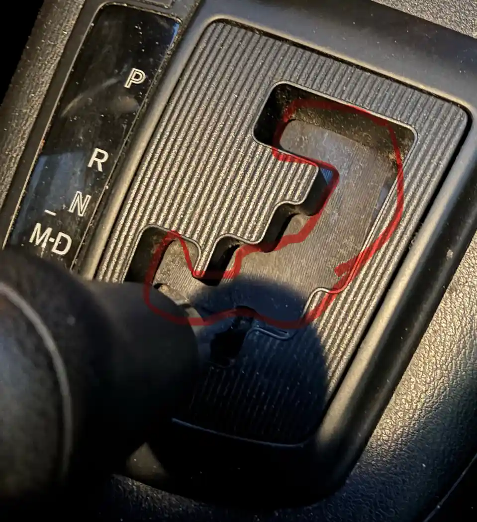 A close-up photo of an automatic car's gear shifter with a red outline drawn around the gear selector, which visually…