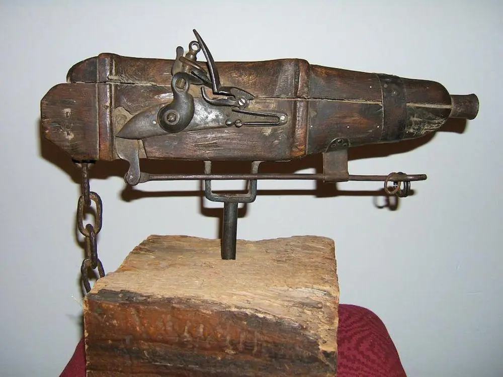A rustic, wooden and metal contraption resembling a firearm mounted on a wooden block, with a chain hanging from its side…
