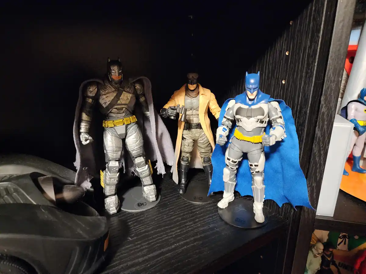 Three Batman action figures in different costumes stand side-by-side on a shelf, with the one in the center wearing a tan…