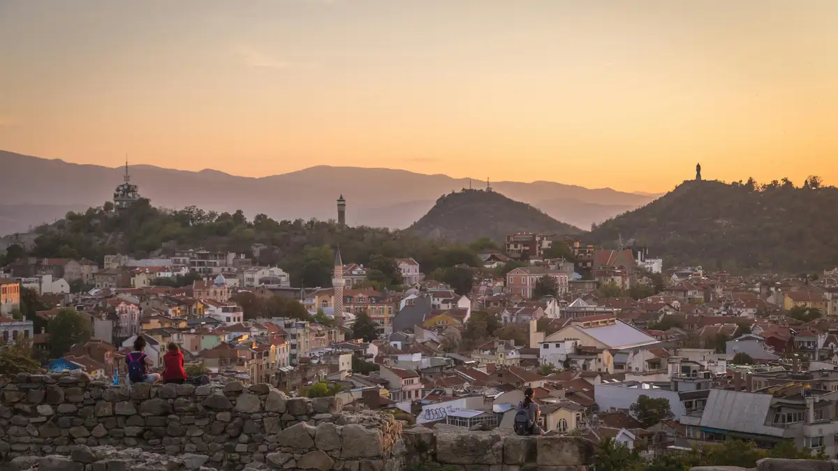 Golden hues engulf ancient ruins, worn smooth by time, as the distant Sredna Gora mountains rise majestically into the fiery…