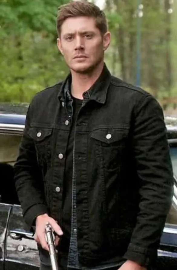 A man with short brown hair, wearing a black denim jacket over a dark shirt, stands outdoors next to a car holding a silver…