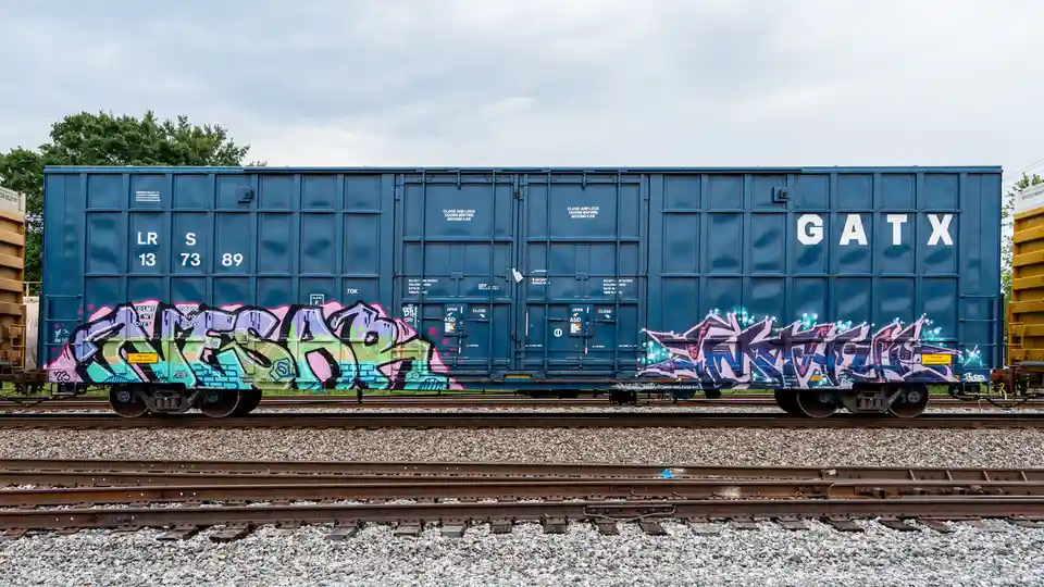 A blue GATX railcar sits on tracks with vibrant graffiti on its sides, featuring colorful stylized lettering and tags. The…