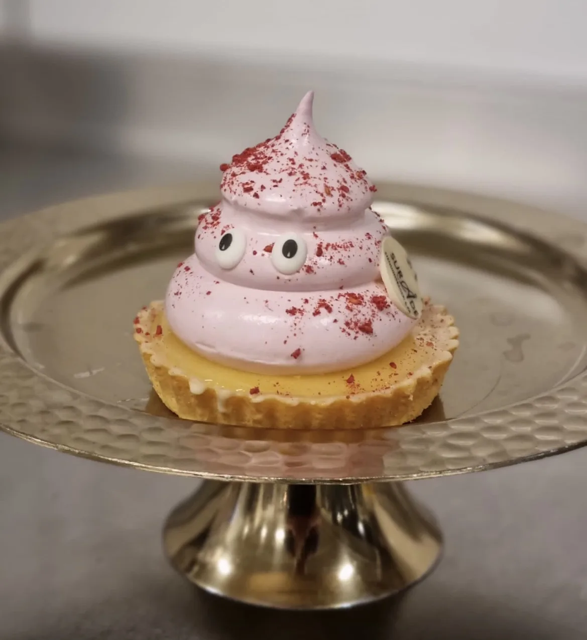 a small pastry tart with a yellow custard base sits on a gold cake stand. The top is a swirl of pink meringue shaped like a…