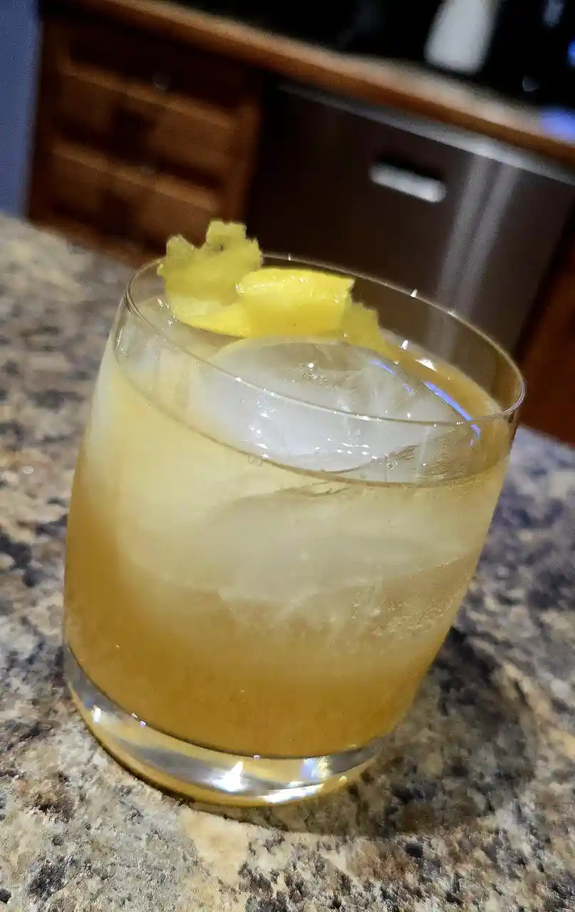 A close-up photo shows a glass of pale yellow cocktail with a large ice cube and pineapple garnish, sitting on a speckled…