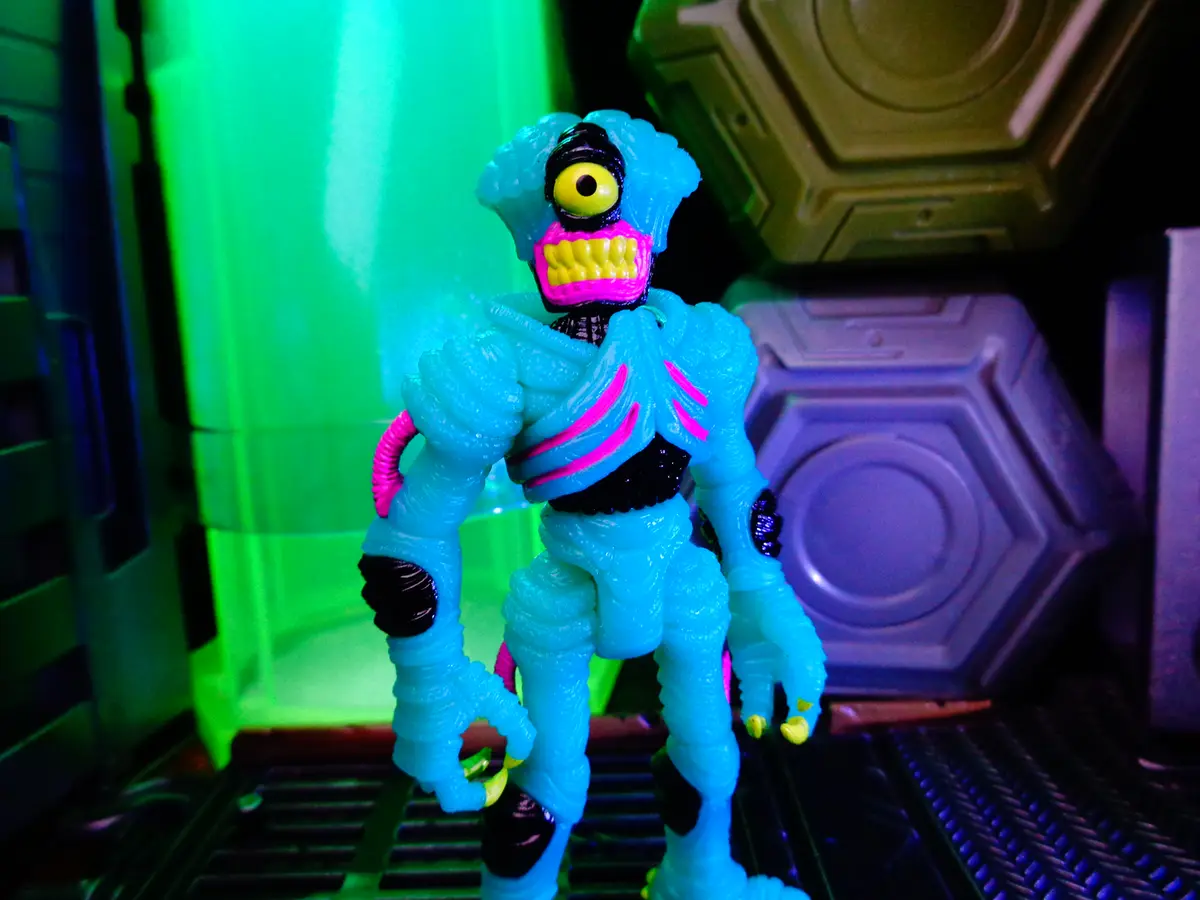 A blue, textured action figure with a single large yellow eye, pink mouth with yellow teeth, and pink stripes on its torso…