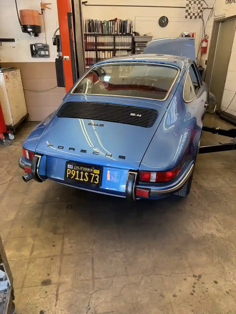 A rear view of a blue 1973 Porsche 911S with its hood open, parked in a garage. The California license plate reads 'P911S…