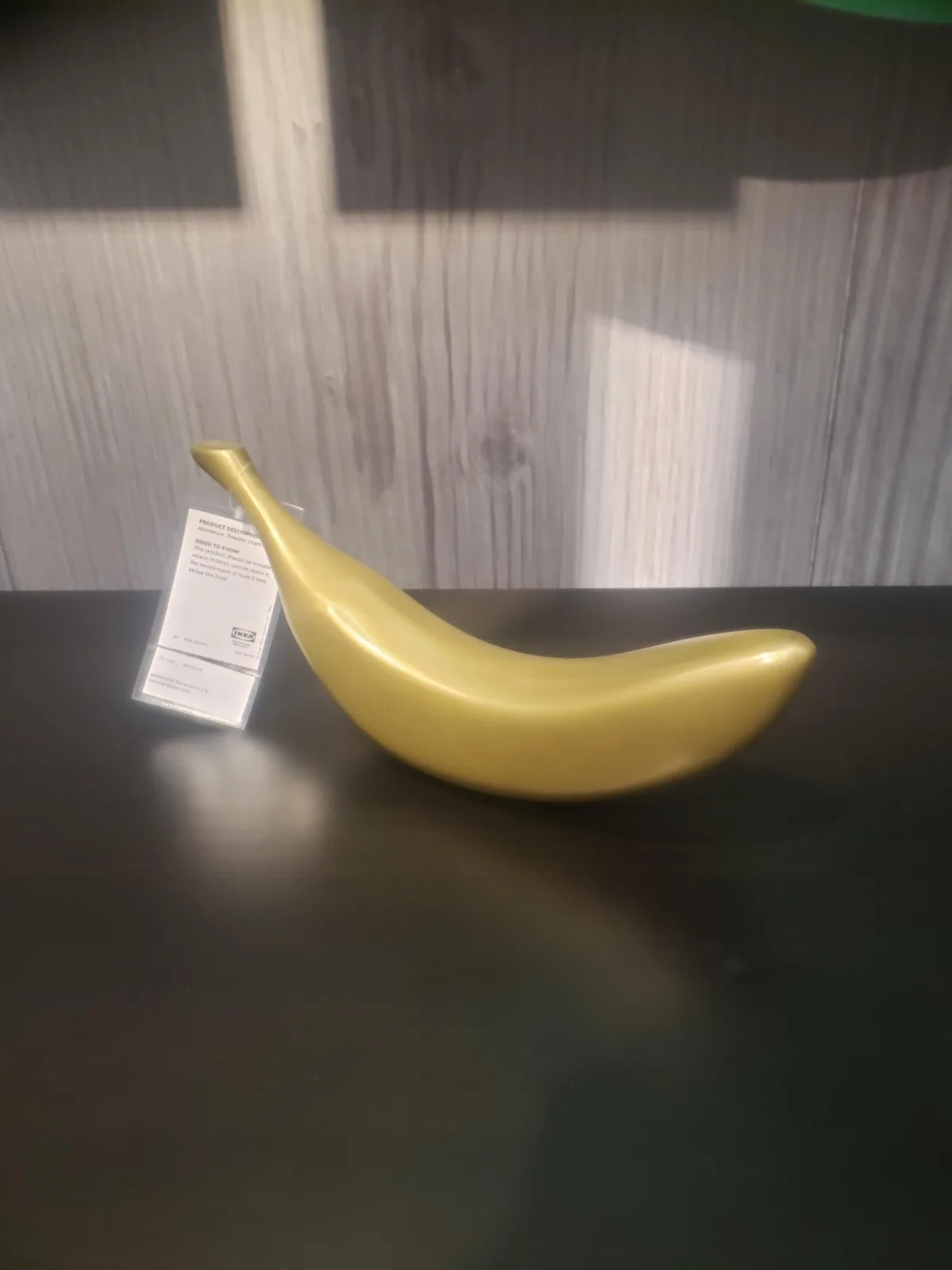 A shiny, gold-colored decorative banana sculpture sits on a dark surface with an IKEA product tag attached. The tag includes…