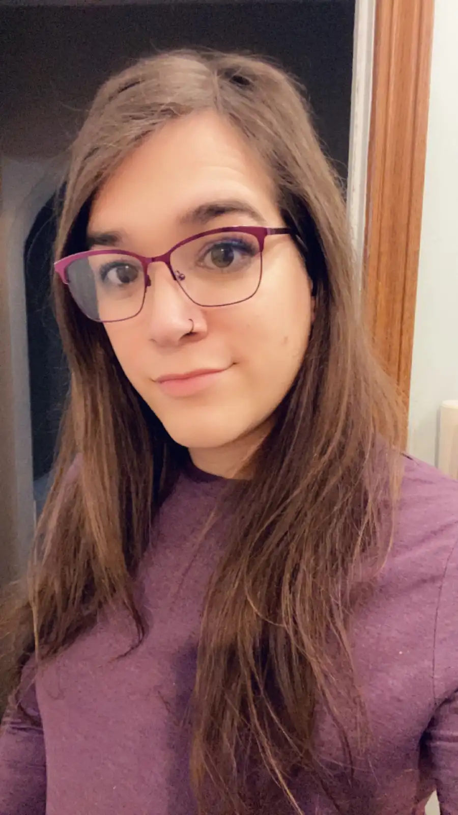 A person with long brown hair, wearing purple-framed glasses and a purple shirt, looks directly at the camera with a slight…
