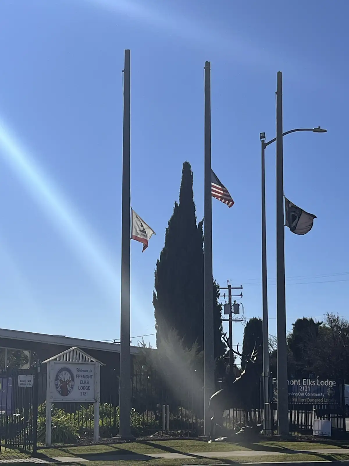 Three flagpoles stand outside Fremont Elks Lodge 2121, with the American flag, California state flag, and a third flag all…