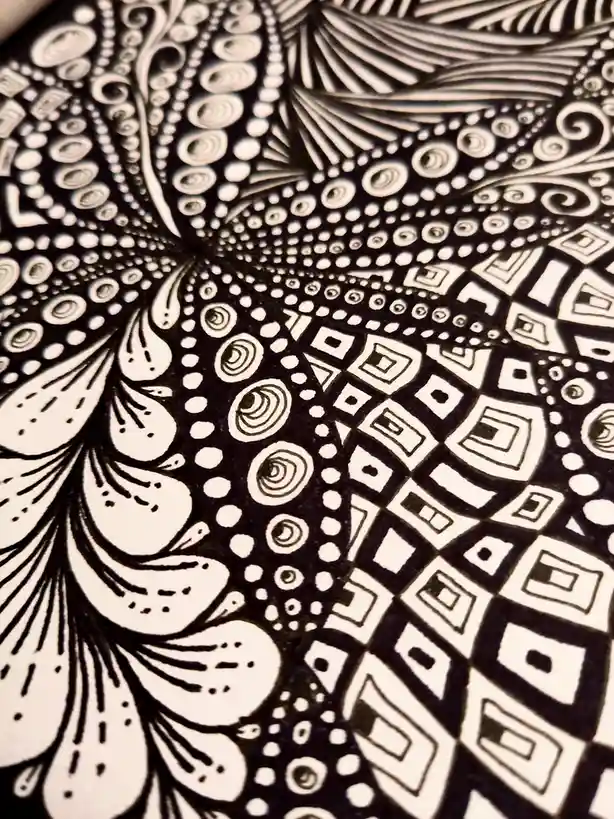 A close-up of intricate black-and-white zentangle art featuring swirling patterns, dots, and geometric shapes like squares…