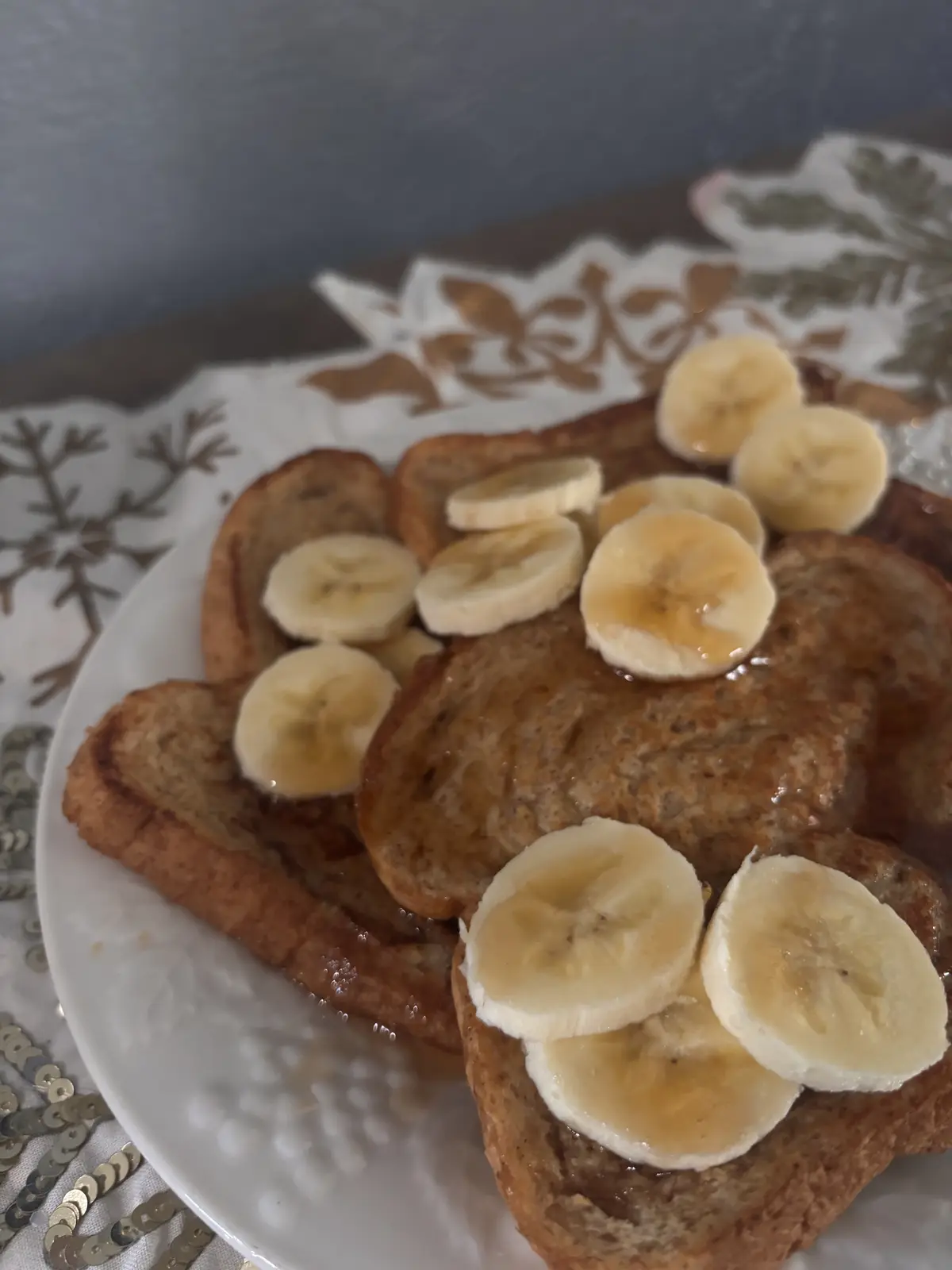 A plate of golden-brown French toast topped with numerous sliced bananas and drizzled with syrup, set on a white plate over…