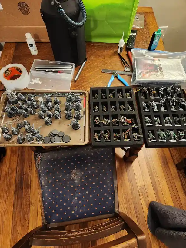 An overhead shot of a cluttered wooden table with three trays of miniature figurines; the left tray holds unpainted grey…