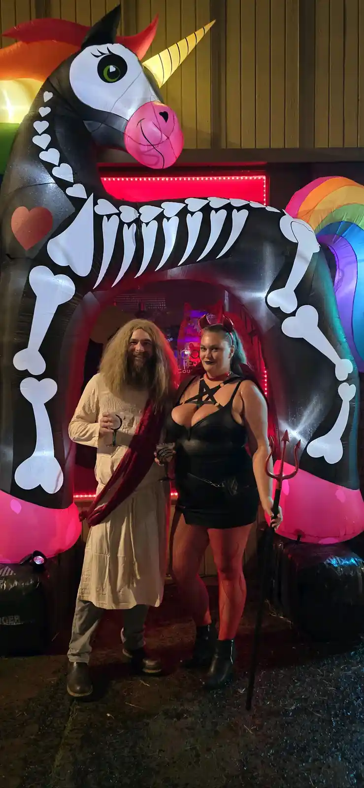 Two people pose in front of a large inflatable skeleton unicorn with a rainbow tail and pink hooves. The man on the left…