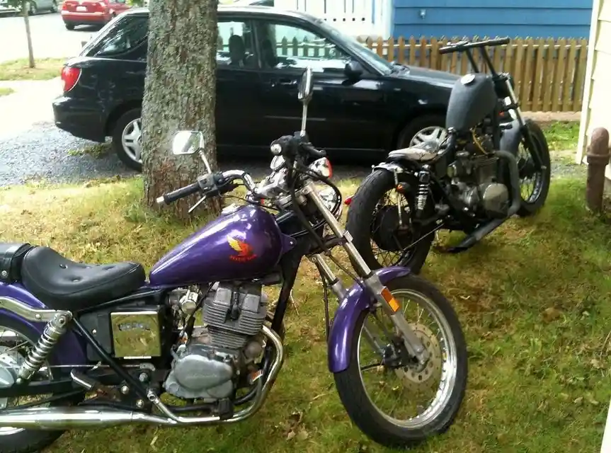 Two motorcycles are parked on grass next to a tree and a black car. The motorcycle in the foreground is purple with a Honda…