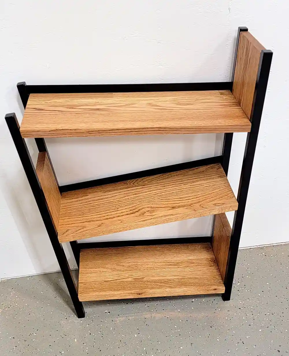 A three-tiered wooden shelf with black metal frame stands against a white wall on a speckled gray floor. The shelves are…